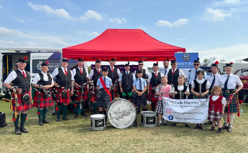 The full band with young Highland dancers in front of a red gazebo at a summer show